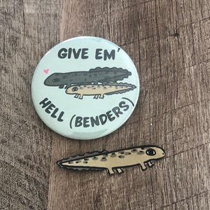 May include: A light blue button with the text "GIVE EM' HELL (BENDERS)" and two salamander illustrations. A separate salamander sticker is also present, featuring a tan body with black spots.