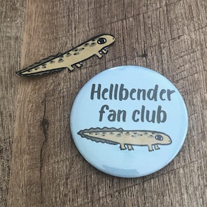 May include: A light blue button with the text "Hellbender fan club" and a cartoon hellbender illustration. A matching hellbender pin is also present. Both items are on a wooden surface.