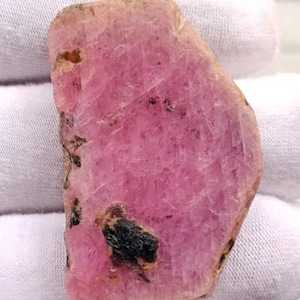 Könnte beinhalten: Ein grob geschnittener, rosa Edelstein mit einer leicht unregelmäßigen Form. Der Stein hat eine lebendige, rosige Farbe mit dunkleren Einschlüssen und einer strukturierten Oberfläche. Der Edelstein wird vor einem weißen Hintergrund präsentiert, um seine natürliche Schönheit hervorzuheben.