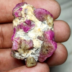 May include: A raw, unpolished mineral specimen featuring vibrant magenta and deep pink crystalline clusters embedded within a rough, textured matrix of off-white and grey stone. The irregular, jagged rock displays a mix of opaque and translucent sections.