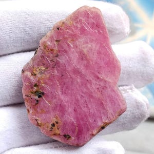 May include: A roughly cut, pink gemstone with a textured surface. The stone has a vibrant, rosy hue with darker flecks and veins running through it. The irregular shape and natural texture suggest it is a raw, unpolished mineral specimen.