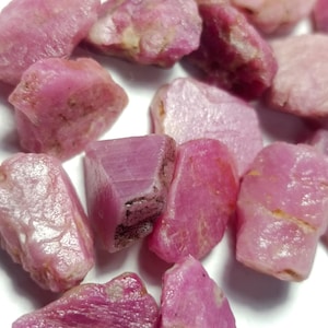 Peut inclure: Une collection de pierres précieuses rubis roses brutes et non polies aux formes irrégulières et aux textures variées. Les pierres présentent une gamme de teintes roses opaques à translucides, avec des facettes naturelles et des inclusions.