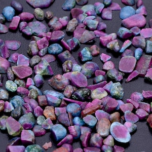 May include: A scattering of raw gemstones in shades of purple, blue, and brown. The stones vary in size and shape, with a mix of smooth and rough textures. The gemstones are scattered across a dark surface, creating a visually appealing display.