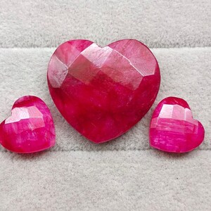 May include: Three faceted, heart-shaped gemstones in varying sizes. The largest is centered, flanked by two smaller hearts. All are a vibrant, deep pink color, with a geometric, cut surface.