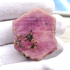Puede incluir: Una gema de rubí rosa en bruto con una estructura cristalina visible. El rubí muestra un color rosa intenso con inclusiones rosas y negras más oscuras. La gema se sostiene en una mano enguantada de blanco.