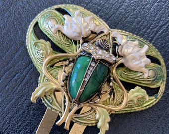 May include: A gold hair clip with a green beetle design. The beetle has a green stone body and is surrounded by gold and green floral details. The clip has two long gold prongs.