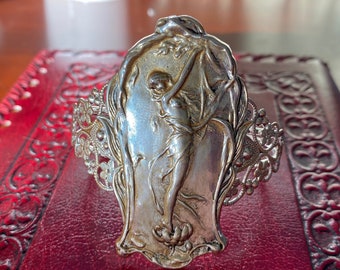 May include: A silver bracelet with a detailed design of a woman standing in a floral setting. The bracelet is made of filigree and has a delicate, feminine look.