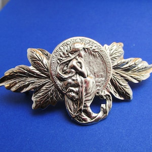 Op de afbeelding: Zilverkleurige haarspeld met een gedetailleerd ontwerp van een vrouw in een cirkel omringd door bladeren.