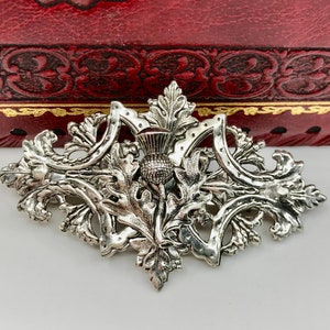 Könnte beinhalten: Eine silberfarbene Brosche mit einem detaillierten Design einer Distelblüte mit Blättern. Die Brosche ist verziert und hat eine Vintage-Ästhetik.