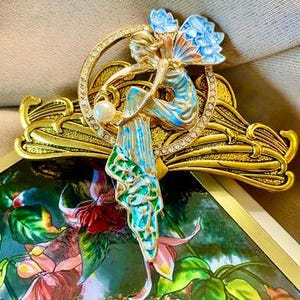 May include: A gold-toned Art Nouveau style brooch featuring a figure with blue wings and a flowing gown, holding a pearl. The brooch is adorned with rhinestones and rests on a decorative gold base. The figure is painted with blue, green, and gold enamel.
