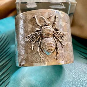 Può includere: Bracciale a polsino argentato con un dettagliato motivo a forma di ape. Il bracciale presenta un motivo geometrico e una superficie strutturata. L'ape è finemente progettata con ali e segmenti del corpo visibili.