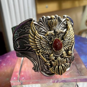 May include: A wide cuff bracelet featuring a silver-toned metal base with intricate scrollwork. The centerpiece is a gold-toned double-headed eagle emblem topped with a crown and accented by a single, smooth, oval-shaped dark red stone at its center.