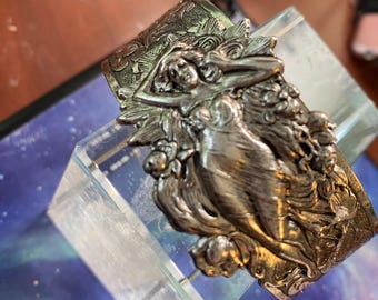 Op de afbeelding: Zilverkleurige manchetarmband met een gedetailleerd reliëf van een liggende vrouw te midden van bloemmotieven. De armband wordt gepresenteerd op een heldere, schuine standaard. De manchet heeft een sierlijke, vintage esthetiek.