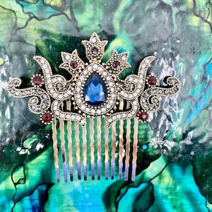 May include: A gold hair comb with a decorative design featuring a large blue gemstone surrounded by smaller clear and red gemstones. The comb has a traditional ornate design.