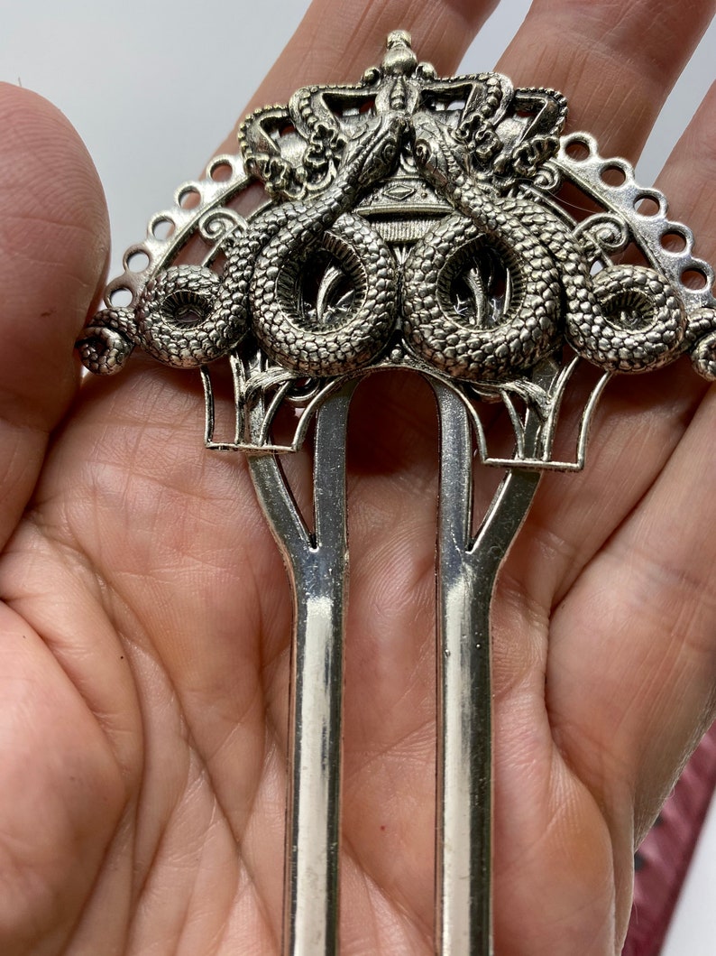 May include: Silver hair fork with a decorative design of two snakes intertwined around a crown. The fork has two prongs.