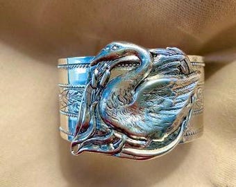 May include: A silver napkin ring with a detailed relief of a bird, possibly a heron or crane, with its head and neck curved. The ring has a decorative band around the top and bottom. The bird's feathers and beak are clearly defined.