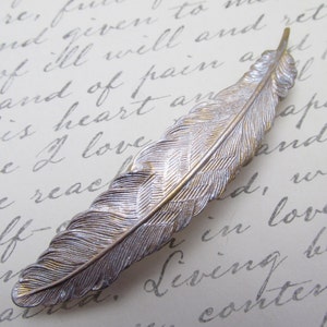 May include: A silver and gold feather-shaped hair clip. The feather has a detailed design and is a delicate accessory.