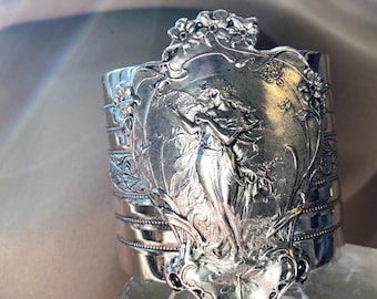 Op de afbeelding: Zilverkleurige manchetarmband met een Art Nouveau-ontwerp. De armband heeft een centraal paneel met een gedetailleerd reliëf van een vrouw en bloemmotieven. De manchet heeft een brede band met decoratieve horizontale lijnen en bloemaccenten.