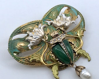 May include: A green and gold jeweled beetle brooch with a pearl pendant. The beetle is set in a green stone with rhinestones and surrounded by gold filigree and white flowers.