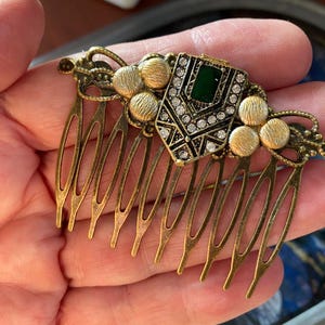 May include: A gold hair comb with a decorative green and clear rhinestone design. The comb has a series of teeth on the bottom.