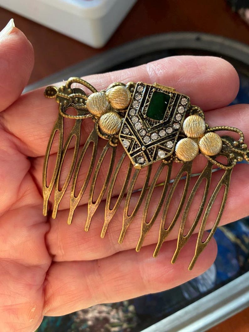May include: A gold hair comb with a decorative green and clear rhinestone design. The comb has a series of teeth on the bottom.
