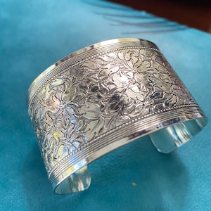 May include: Silver cuff bracelet with an embossed floral design. The wide bracelet features detailed, raised floral patterns across its surface. The cuff has a polished finish and a slight curve to fit the wrist. The background is a teal color.