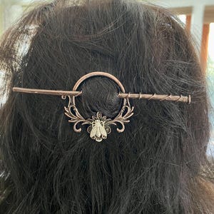 Pode incluir: Grampo de cabelo cor de cobre com um design circular e um motivo decorativo de abelha. O grampo está inserido em cabelo castanho escuro, destacando os seus detalhes intrincados. Este acessório de cabelo adiciona um toque de elegância.