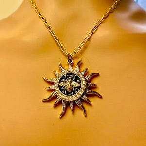 Könnte beinhalten: Goldfarbene Halskette mit einem Sonnenstrahlen-Anhänger mit einem zentralen Bienen-Design. Der Anhänger ist mit klaren Kristallen und einem schwarzen Hintergrund verziert. Die Kette ist im einfachen Kettenglied-Stil gehalten.