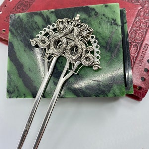 May include: Silver hair stick with a decorative top featuring two intertwined snakes with a crown. The snakes are detailed with scales and the crown has a decorative design.