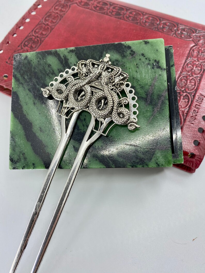 May include: Silver hair stick with a decorative top featuring two intertwined snakes with a crown. The snakes are detailed with scales and the crown has a decorative design.