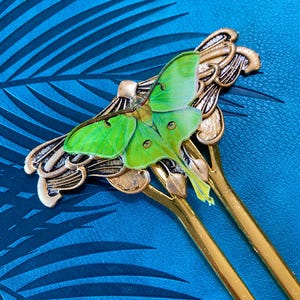 May include: A gold-coloured hair stick with a green and yellow moth design. The moth has detailed wing patterns. The hair accessory is set against a textured blue background with a palm leaf pattern.