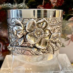 Puede incluir: Un recipiente cilíndrico plateado con un diseño floral en relieve. El recipiente presenta una flor grande y detallada en el centro, rodeada de hojas y patrones intrincados. El recipiente es probablemente de metal.
