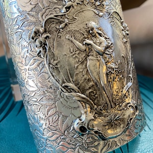 Könnte beinhalten: Silberfarbenes Armreif-Armband mit Jugendstil-Design. Das Armband zeigt ein zentrales Relief einer Frau, umgeben von floralen und blattartigen Mustern. Der Armreif ist breit und hat eine detaillierte, geprägte Textur, die eine Vintage-Ästhetik erzeugt.