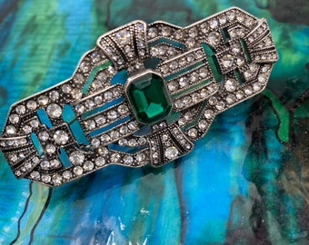 May include: Silver-toned Art Deco brooch with a rectangular emerald-green center stone, surrounded by clear, sparkling rhinestones. The brooch has an intricate geometric design, perfect for adding a touch of vintage glamour to any outfit.