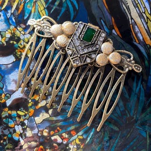 May include: A gold hair comb with a decorative design featuring a green and clear rhinestone embellishment. The comb has a series of teeth on the bottom.