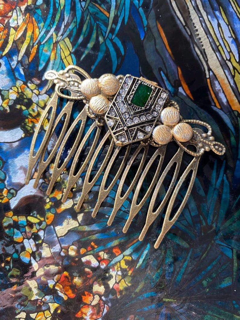 May include: A gold hair comb with a decorative design featuring a green and clear rhinestone embellishment. The comb has a series of teeth on the bottom.