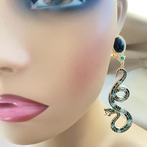 May include: Gold-tone snake earrings with black and green enamel detailing and clear crystal accents. The earrings feature a black oval stone and a small green stone at the top. The snake design is detailed and elegant.