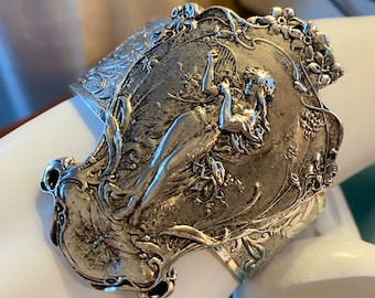 May include: A silver-toned cuff bracelet with an ornate design featuring a fairy figure with wings and floral accents.