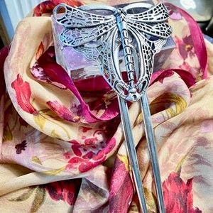 May include: A silver-toned metal hair fork featuring a detailed dragonfly design with intricate cut-out wing patterns. The dragonfly body is elongated and textured, extending into two long, smooth prongs designed for securing hair in place.
