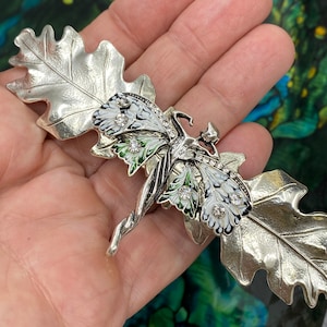 May include: A silver-toned brooch featuring a fairy with wings and a floral design. The fairy is surrounded by two oak leaves with a white and green enamel finish. The brooch is adorned with clear rhinestones.