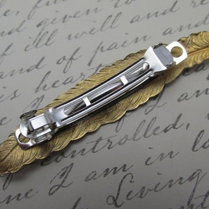 May include: Gold feather hair clip with a silver metal clasp. The feather is detailed with a textured design.