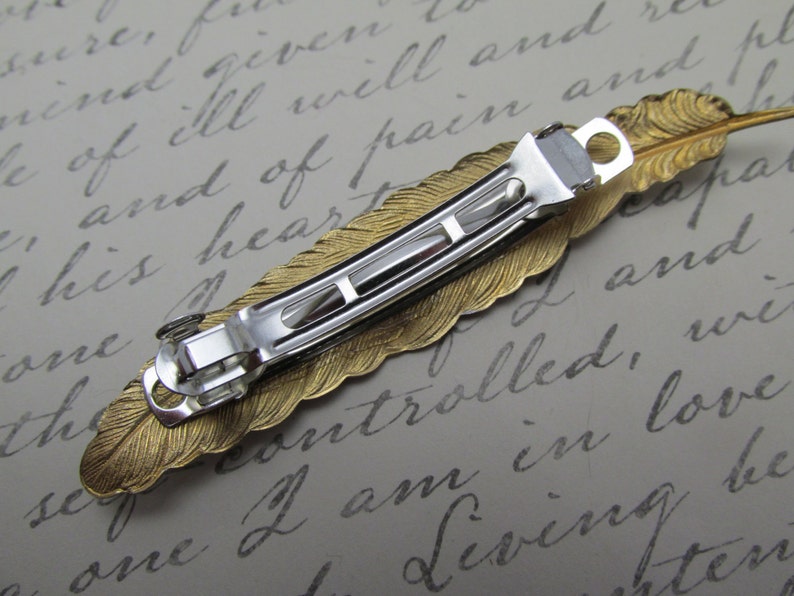 May include: Gold feather hair clip with a silver metal clasp. The feather is detailed with a textured design.