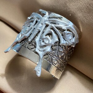 Puede incluir: Brazalete de puño plateado con un diseño floral y de volutas ornamentado. El puño presenta una superposición texturizada en relieve con una forma orgánica y fluida. El diseño del brazalete incluye patrones florales detallados.