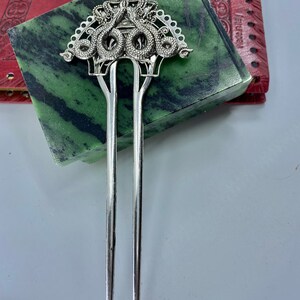 May include: Silver hair stick with a decorative top featuring a crown and three intertwined snakes. The snakes are detailed with scales and have a stylized design.