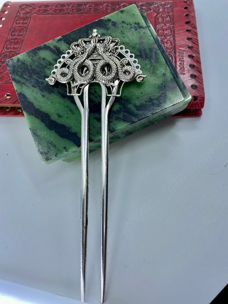 May include: Silver hair stick with a decorative top featuring a crown and three intertwined snakes. The snakes are detailed with scales and have a stylized design.