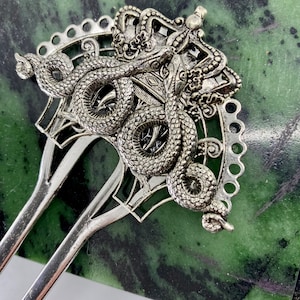 May include: Silver hair comb with a decorative design of a crown and two snakes. The snakes are intertwined and the crown is ornate.
