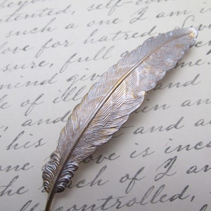 May include: A silver feather-shaped hair clip with a textured design. The clip is on a white surface with handwritten text.