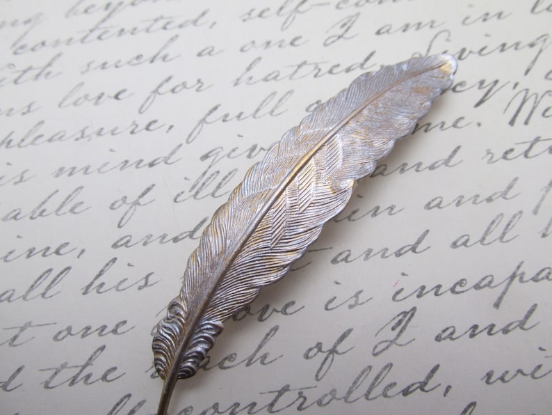 May include: A silver feather-shaped hair clip with a textured design. The clip is on a white surface with handwritten text.