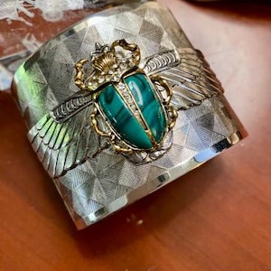 May include: Silver cuff bracelet with a geometric pattern. The bracelet features a gold-toned scarab beetle with a green malachite stone and crystal accents. The beetle has wings and a crown. The cuff is wide and has a textured surface.
