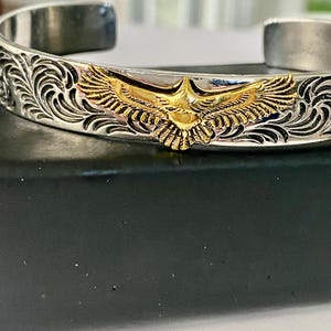 May include: Silver bracelet with a gold eagle design. The bracelet has an intricate floral pattern.